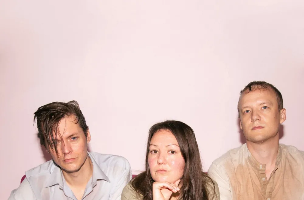 Three people sit on a red sofa and look into the camera.