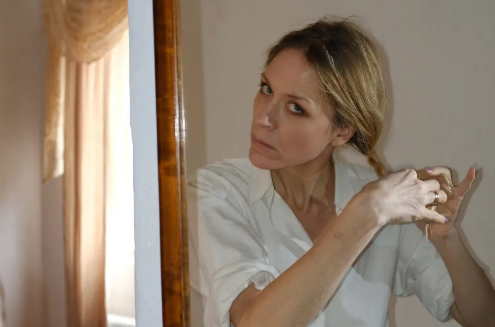 Blonde woman in a white shirt braids her hair while looking at herself in a mirror.